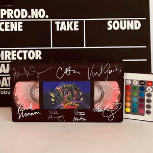 May include: A black and white clapperboard with the words "PROD.NO., SCENE, TAKE, SOUND, DIRECTOR, CAM, DAT, PRODU" printed on it. A black VHS tape with a red light inside is sitting on a white surface. The tape has a picture of the movie "Little Shop of Horrors" on it. The tape is signed by the cast and crew. A remote control with colored buttons is sitting next to the tape.