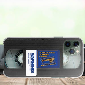 May include: A black and clear phone case with a design that looks like a VHS tape. The case has a blue sticker that says "WARNING! If this label is broken, you must purchase this video. To PLAY is human. TO REWIND is divine! BLOCKBUSTER VIDEO WOW! What a Difference!"