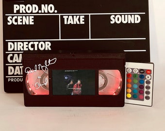 May include: A red and black VHS tape with a movie poster inside. The tape has a red light on the side and a remote control with color buttons. The tape is signed by Phil Noftsinger.