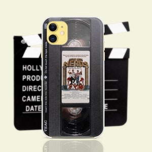 Puede incluir: Una funda para teléfono negra y gris con un diseño de cinta VHS. La funda presenta un póster de película para la película "La venganza de los nerds" con el texto "Inserte este lado en la grabadora No toque la cinta en el interior" y "TEAC" impreso en el lateral.