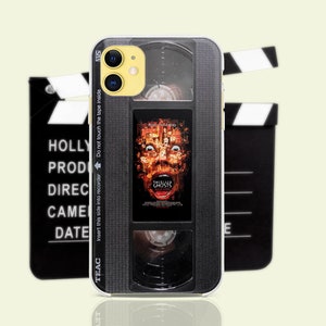 May include: A black and gray phone case with a VHS tape design. The case features a movie poster for the film "The Ghostly" with a scary face on it. The case has a TEAC logo and the words "Insert this side into recorder Do not touch the tape inside".
