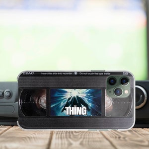 May include: A black and green iPhone case with a VHS tape design featuring the movie "The Thing". The case has a clear back and a black front with the text "Insert this side into recorder Do not touch the tape inside" and "VHS".