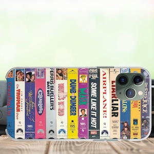 May include: Clear phone case featuring a colorful design of VHS tapes. The tapes include movie titles like "Groundhog Day," "Clueless," and "Dumb and Dumber." The case has a clear border and covers the back of a smartphone.