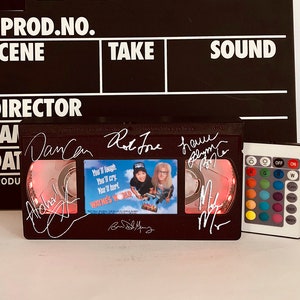 May include: A black VHS tape with a red light shining through the clear plastic window. The tape has the title "Home Alone" and is signed by the actors and director. The tape is next to a black remote control with colored buttons.