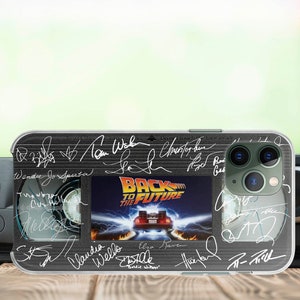 May include: A clear phone case featuring a black and white image of a VHS tape with the text "Back to the Future" and signatures of the cast and crew. The case has a green camera lens.