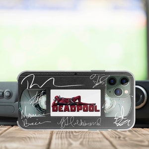 May include: A black and clear phone case with a Deadpool movie theme. The case features a black and white image of Deadpool lying down with the word "DEADPOOL" in white letters. The case also has several signatures in white ink.