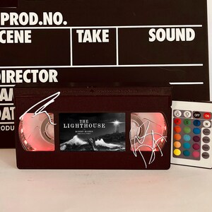 May include: A black and red VHS tape with a white label that reads "The Lighthouse" and a red light shining through the tape. The tape is sitting on a white surface next to a black and white remote control with a variety of colored buttons.