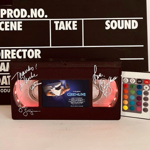 May include: A black VHS tape with a red light shining through the clear plastic window. The tape is signed by the director and the cast of the movie "Gremlins". The tape is on a black surface with a white clapperboard behind it. The clapperboard has the words "Prod. No.", "Scene", "Take", "Sound", and "Director" written on it. There is a remote control with colored buttons on the right side of the image.