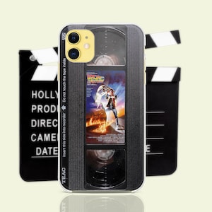 Puede incluir: Una funda para teléfono en blanco y negro con un diseño de cinta VHS vintage. La funda presenta una ventana de plástico transparente con un póster de película para "Regreso al futuro" y el texto "TEAC" e "Inserte este lado en la grabadora No toque la cinta en el interior".