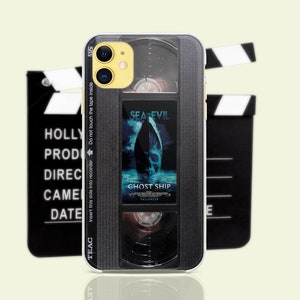 May include: A black and silver phone case with a VHS tape design. The case features a movie poster for the film "Sea Evil: Ghost Ship" with the text "Insert this side into recorder Do not touch the tape inside" and "TEAC" printed on the side.