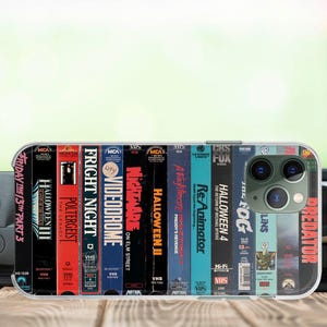 Op de afbeelding: Een transparante telefoonhoes met een collage van vintage VHS-filmhoezen, waaronder titels als "Friday the 13th Part 3", "Halloween II" en "Predator". De hoes heeft een glanzende afwerking en een modern smartphone-ontwerp.