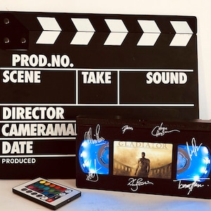 May include: A black and white clapperboard with the words "PROD. NO., SCENE, TAKE, SOUND, DIRECTOR, CAMERAMAN, DATE, PRODUCED" printed on it. Below the clapperboard is a black VHS tape with a blue light shining through the window. The tape has the title "GLADIATOR" printed on it and several signatures. The tape is accompanied by a remote control with colored buttons.