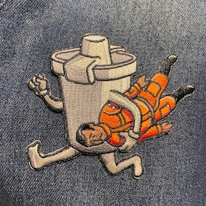 May include: A white and gray embroidered patch of a cartoon shaker with a person in an orange jumpsuit trapped inside. The shaker has legs and arms and is running. The person is upside down and has their arms and legs outstretched.
