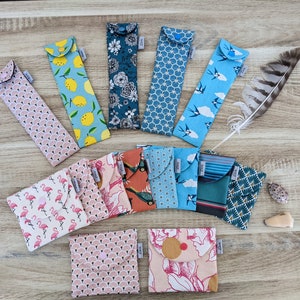 Peut inclure: Une collection de 13 pochettes en tissu colorées avec différents motifs, notamment des flamants roses, des citrons, des oiseaux et des motifs géométriques. Les pochettes sont toutes fermées par un bouton et sont disposées en demi-cercle sur une surface en bois.