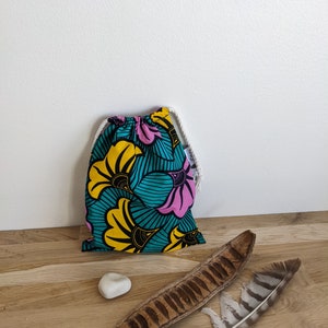 Peut inclure: Un sac à cordon avec un motif floral vibrant en turquoise, jaune et rose. Le sac est en tissu et possède une fermeture à cordon blanche. Une petite pierre blanche, une gousse de graines et une plume sont sur la surface en bois.