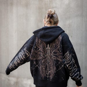 May include: Black hooded sweatshirt with a large, detailed graphic on the back and sleeves. The design features a central sword motif with an eye, surrounded by stylized wings and text. The artwork is in a faded, distressed style.