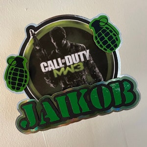 Peut inclure: Un cake topper avec le logo Call of Duty: MW3 et un soldat. Le topper comprend le nom "JAKOB" en vert, bordé de noir, et deux graphiques de grenades vertes. Le design est sur un fond argenté holographique.