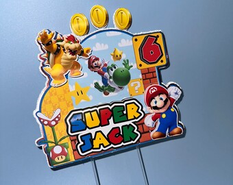 Könnte beinhalten: Ein farbenfroher Tortenaufsatz mit Super Mario-Charakteren. Er zeigt Mario, Yoshi, Bowser und Goldmünzen, zusammen mit der Zahl 6 und dem Namen "SUPER JACK". Der Aufsatz hat klare Kunststoffstäbe zum Einstecken.