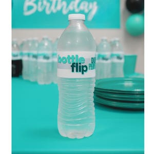 May include: A clear plastic water bottle with a white cap and a label that says "bottle flip". The bottle is on a teal surface, with other bottles and party supplies in the background. The words "DUDE PERFECT Birthday" are visible.