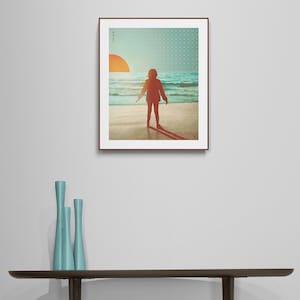 May include: A framed print depicting a silhouette of a person standing on a beach with the sun setting in the background. The print has a blue and green geometric pattern border.