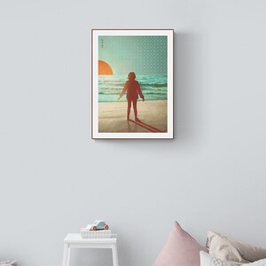 May include: A framed print depicting a person standing on a beach, facing away from the viewer, looking out towards the sea. The sun is setting in the distance, creating a warm glow over the scene. The print has a simple, minimalist style.