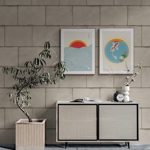 May include: Two framed prints with Japanese-inspired artwork. The print on the left features a red sun setting over a blue ocean with waves. The print on the right features a red and white koi fish swimming in a blue circle with lily pads. The prints are hanging on a light gray brick wall above a white cabinet with a woven rattan front.