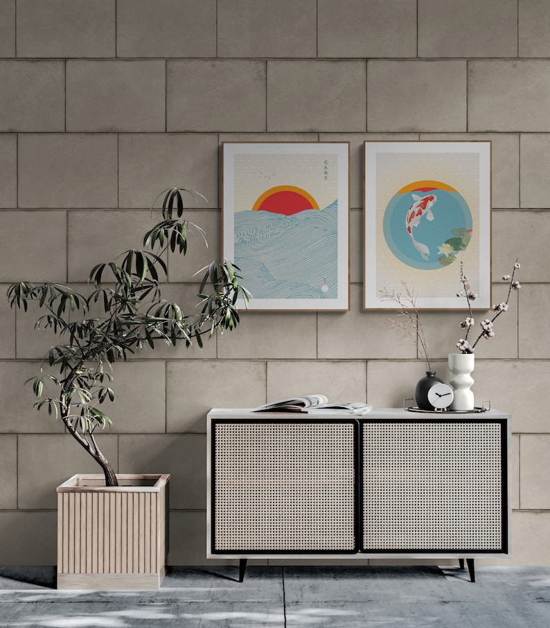May include: Two framed prints with Japanese-inspired artwork. The print on the left features a red sun setting over a blue ocean with waves. The print on the right features a red and white koi fish swimming in a blue circle with lily pads. The prints are hanging on a light gray brick wall above a white cabinet with a woven rattan front.