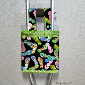 May include: A black fabric crutch bag with a colorful flip-flop pattern. The bag has a green polka dot trim and straps to attach to the crutches. The bag is designed to hold small items and is a practical accessory.