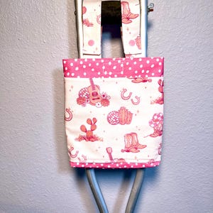 May include: A fabric tote bag with two straps, featuring a white background with a pink western-themed print of cowboy boots, guitars, cacti, horseshoes, and disco balls. The bag has a pink and white polka dot border and the text Carlye Creations.