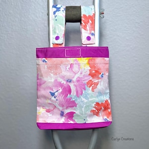 May include: A floral print tote bag with a magenta base and trim, attached to a gray metal frame. The bag features a watercolor design with pink, red, and blue flowers. The top of the bag has a magenta band. The bag is attached to the frame with fabric straps.