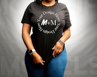 May include: Black t-shirt with a white circular design that reads "Your Design Here" and "M+M". The model is wearing blue jeans and a white smartwatch. The background is a gradient of gray.