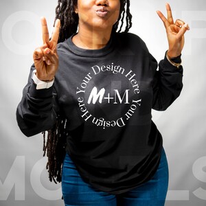 May include: A black long-sleeved shirt with a white circle design that says "Design Your Here Here Your Design M+M". The person wearing the shirt is smiling and giving a peace sign with both hands.
