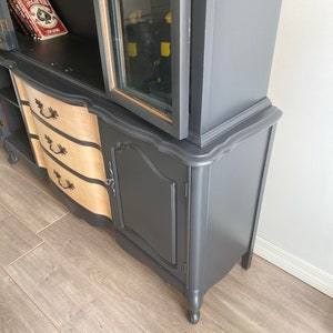 May include: A vintage hutch painted black with a glass door, two drawers, and a single door cabinet. The hutch has a curved top and sits on four legs.