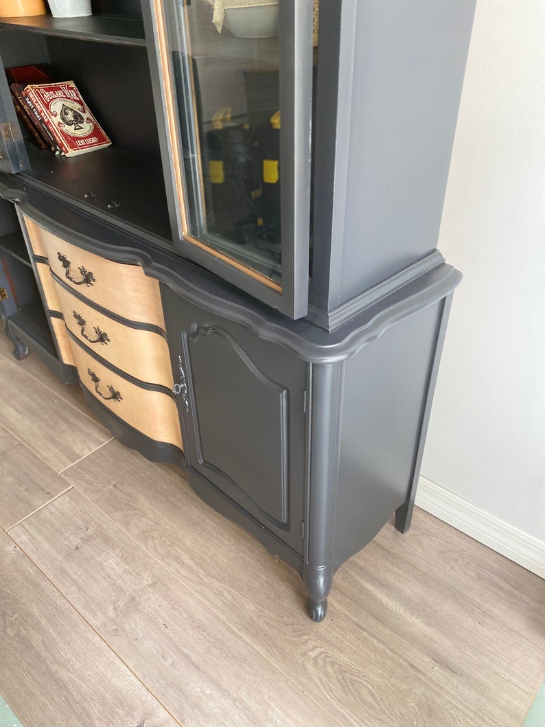 May include: A vintage hutch painted black with a glass door, two drawers, and a single door cabinet. The hutch has a curved top and sits on four legs.