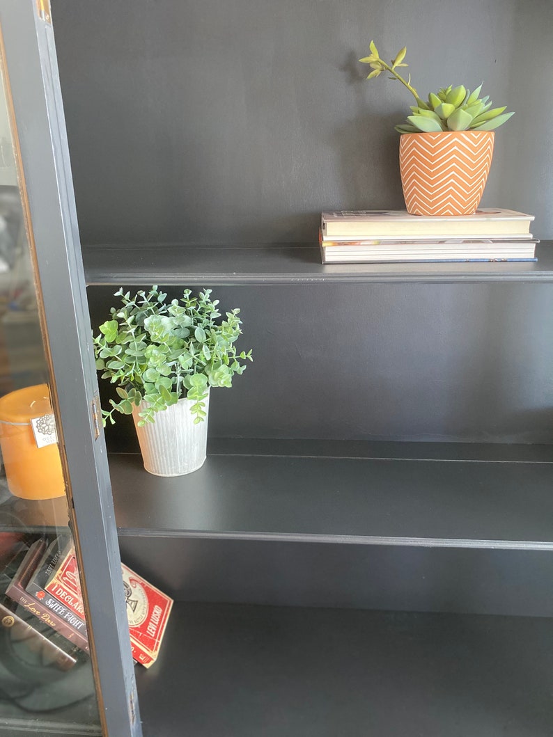 May include: Black wooden bookcase with three shelves. The top shelf has a stack of books and a small potted succulent plant with a brown and white patterned pot. The middle shelf has a small potted plant with green leaves and a white pot. The bottom shelf has a stack of books with red and white covers.