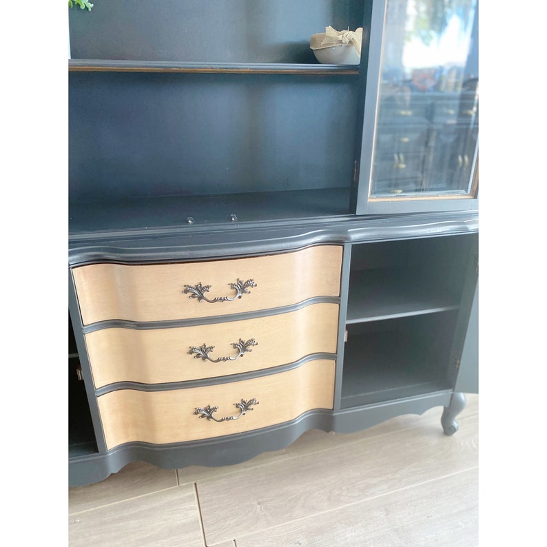 May include: A black painted hutch with three drawers and a glass door. The drawers have a wood veneer front and black handles. The hutch has a shelf inside the glass door.