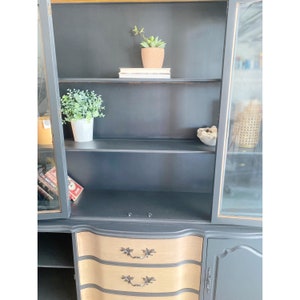 May include: A black hutch with glass doors and two drawers. The hutch has three shelves inside and is decorated with gold accents. The drawers have a wood grain finish and are accented with black handles.