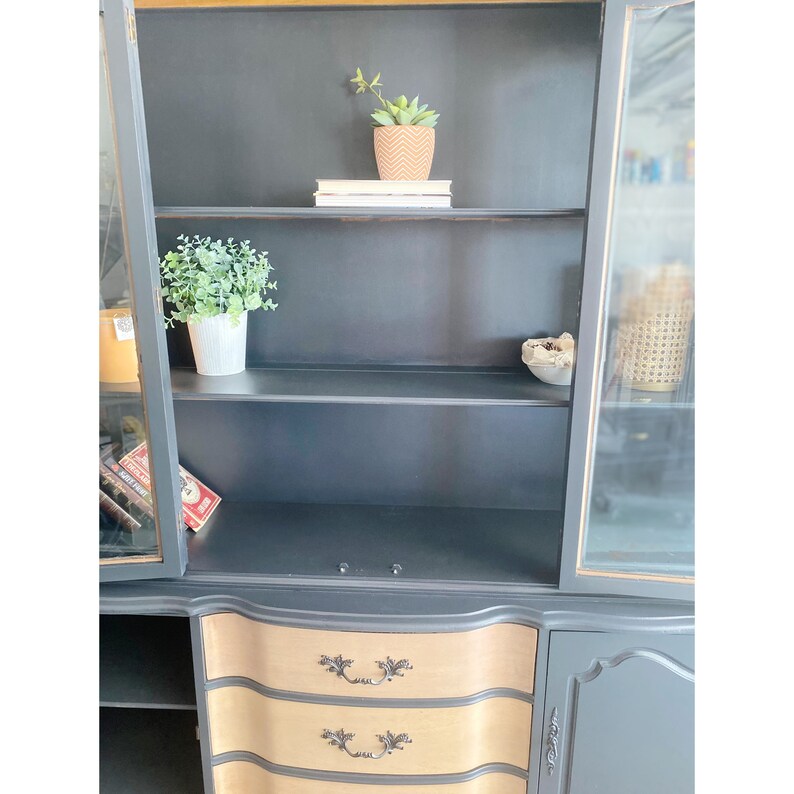 May include: A black hutch with glass doors and two drawers. The hutch has three shelves inside and is decorated with gold accents. The drawers have a wood grain finish and are accented with black handles.