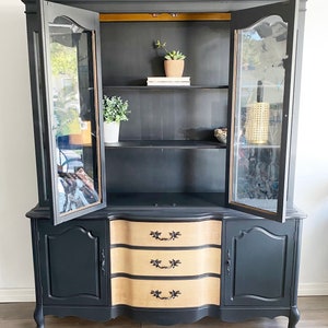 May include: A vintage hutch painted black with glass doors and a wood-toned bottom with three drawers. The hutch has a curved top and bottom and features gold accents.