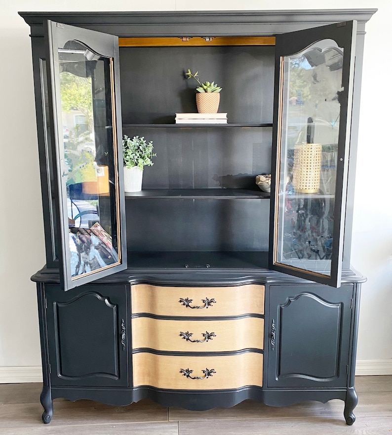 May include: A vintage hutch painted black with glass doors and a wood-toned bottom with three drawers. The hutch has a curved top and bottom and features gold accents.