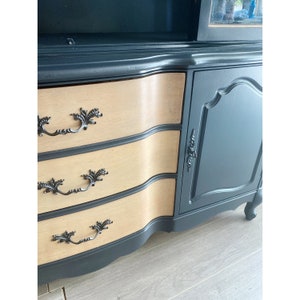 May include: A black painted wooden cabinet with three drawers and a single door. The drawers have a light wood finish and ornate black metal handles. The cabinet has a curved front and sits on four legs.