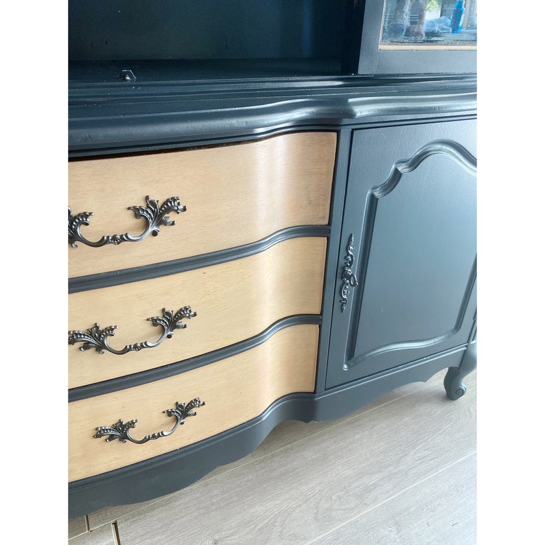 May include: A black painted wooden cabinet with three drawers and a single door. The drawers have a light wood finish and ornate black metal handles. The cabinet has a curved front and sits on four legs.