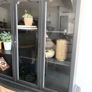 May include: Black painted china cabinet with glass doors and two drawers. The cabinet has a decorative trim around the glass doors and a curved design on the bottom drawers. The cabinet is empty except for a small potted plant on the top shelf and a wicker lamp on the bottom shelf.