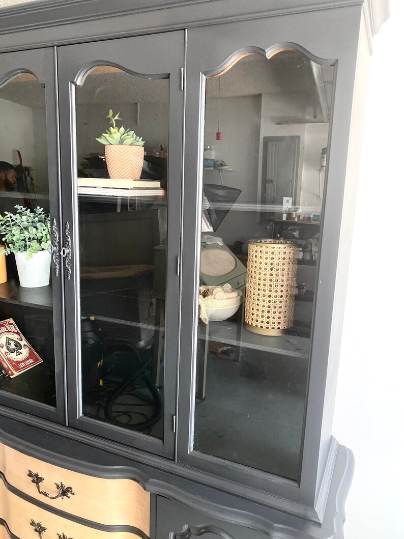 May include: Black painted china cabinet with glass doors and two drawers. The cabinet has a decorative trim around the glass doors and a curved design on the bottom drawers. The cabinet is empty except for a small potted plant on the top shelf and a wicker lamp on the bottom shelf.