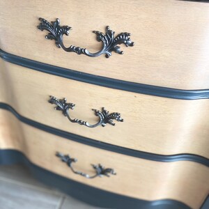 May include: A dresser with three drawers, each with a decorative black metal handle. The drawers are made of light wood and have a dark blue painted finish on the edges.