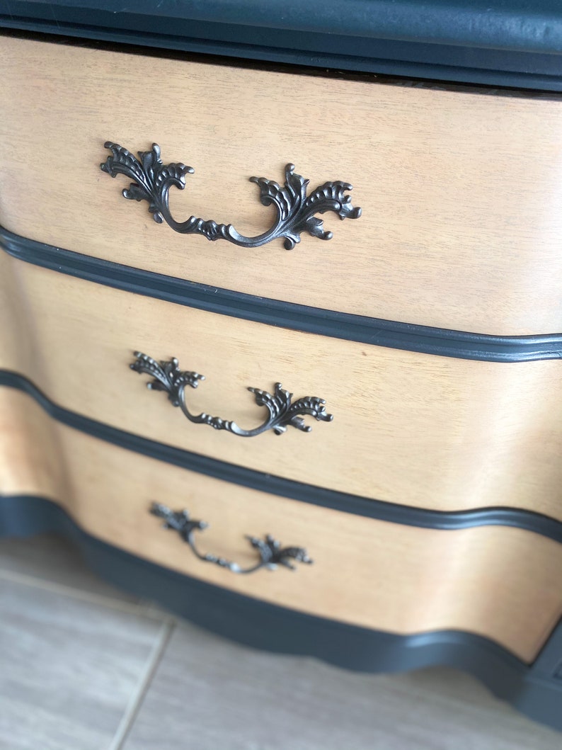 May include: A dresser with three drawers, each with a decorative black metal handle. The drawers are made of light wood and have a dark blue painted finish on the edges.
