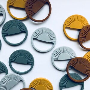 May include: A collection of circular, silicone sun-shaped objects in various colors, including mustard yellow, brown, gray, and teal. Each object has a handle and a sunburst design. The objects are scattered on a white surface.