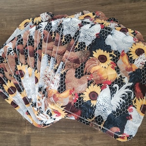 Op de afbeelding: Een stapel stoffen maandverbanden met een boerderijdierenprint. De maandverbanden hebben een herhalend patroon van kippen, hanen en zonnebloemen op een zwarte achtergrond met een kippengaasontwerp. De maandverbanden zijn in verschillende tinten bruin, geel, wit en zwart.
