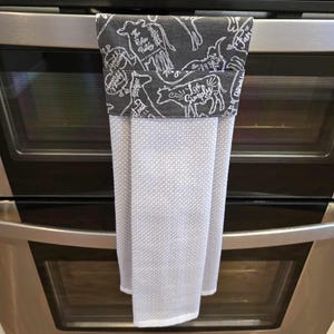 May include: A grey and white kitchen towel with a black and white design of farm animals and the text "The Rustic Pantry" and "Live Simply". The towel is hanging on a stainless steel oven door.