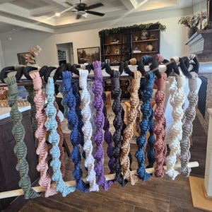 May include: A display of handmade macrame plant hangers in various colors, including green, pink, blue, purple, brown, and white. Each hanger features a spiral design and is suspended from a wooden dowel. The hangers are displayed indoors.